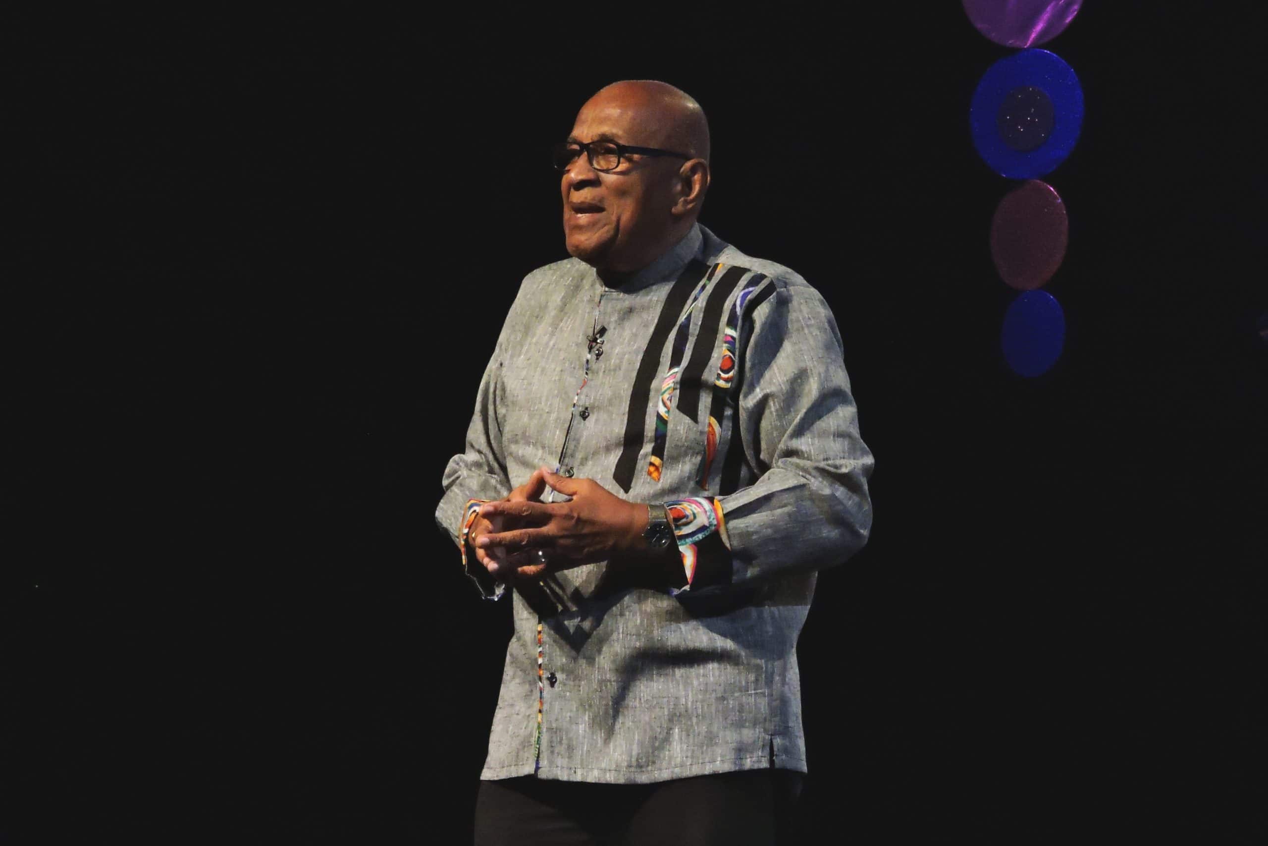 Caribbean poet Paul Keens-Douglas, wearing glasses and a grey patterned shirt, stands on a stage and gestures whilst speaking.