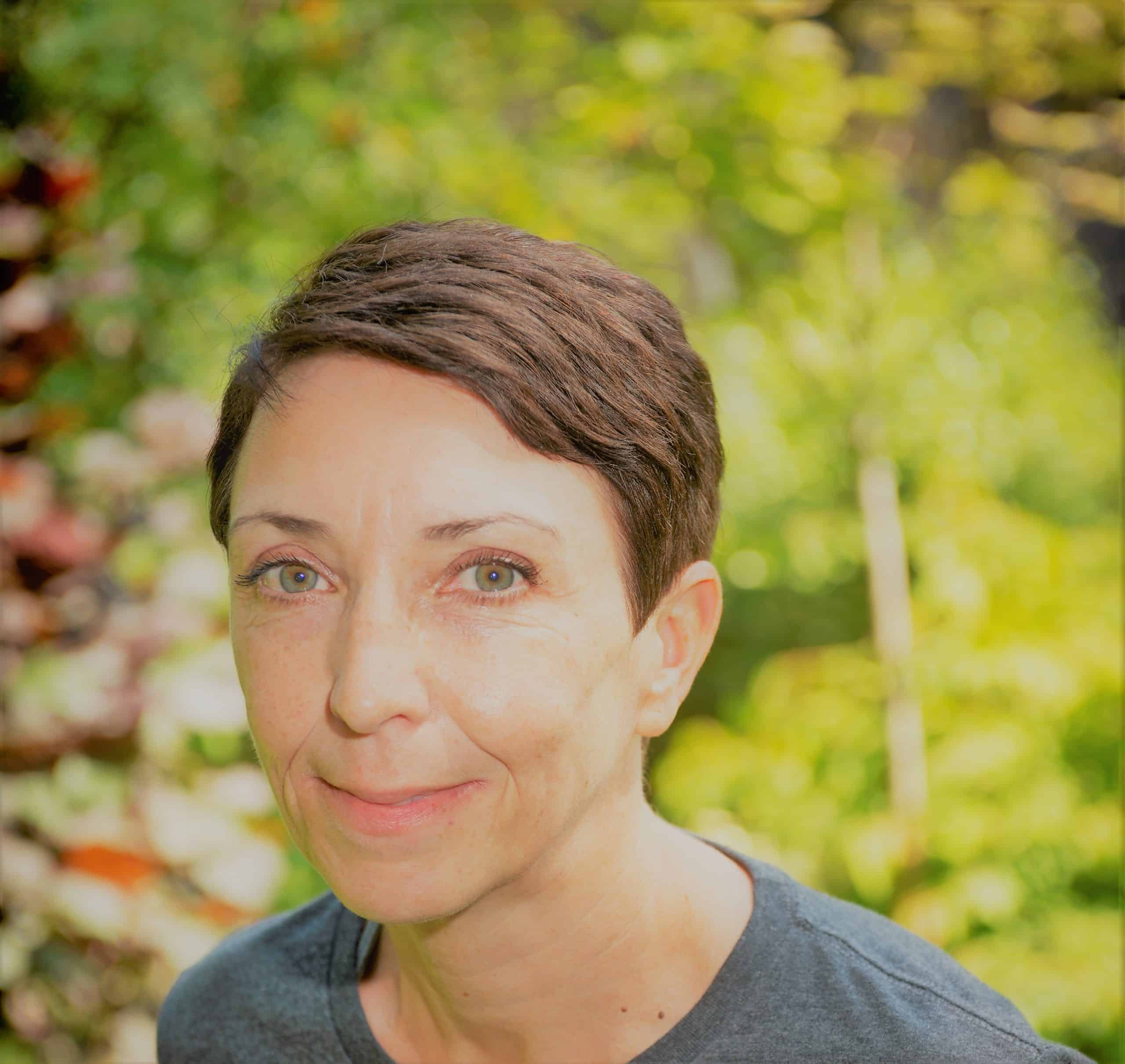 A woman with short brown hair looking towards the camera against a green outdoor background.