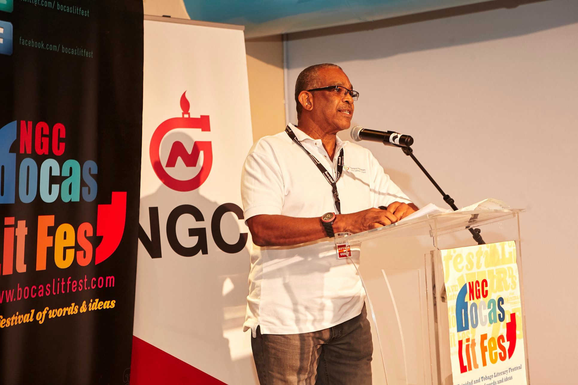 A man in a white polo shirt shares publishing tips at a lectern branded with the NGC Bocas Lit Fest logo.