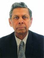 An older man with grey hair wears a dark suit, light shirt, and patterned tie, looking directly at the camera against a plain white background.