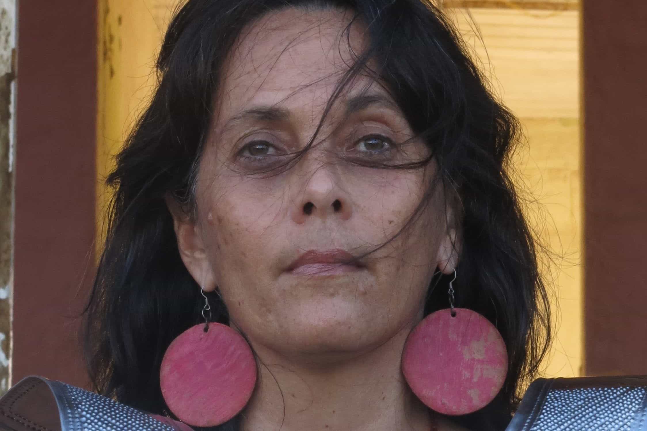 A portrait of contemporary artist Annalee Davis, a woman with dark hair wearing large, circular pink earrings.