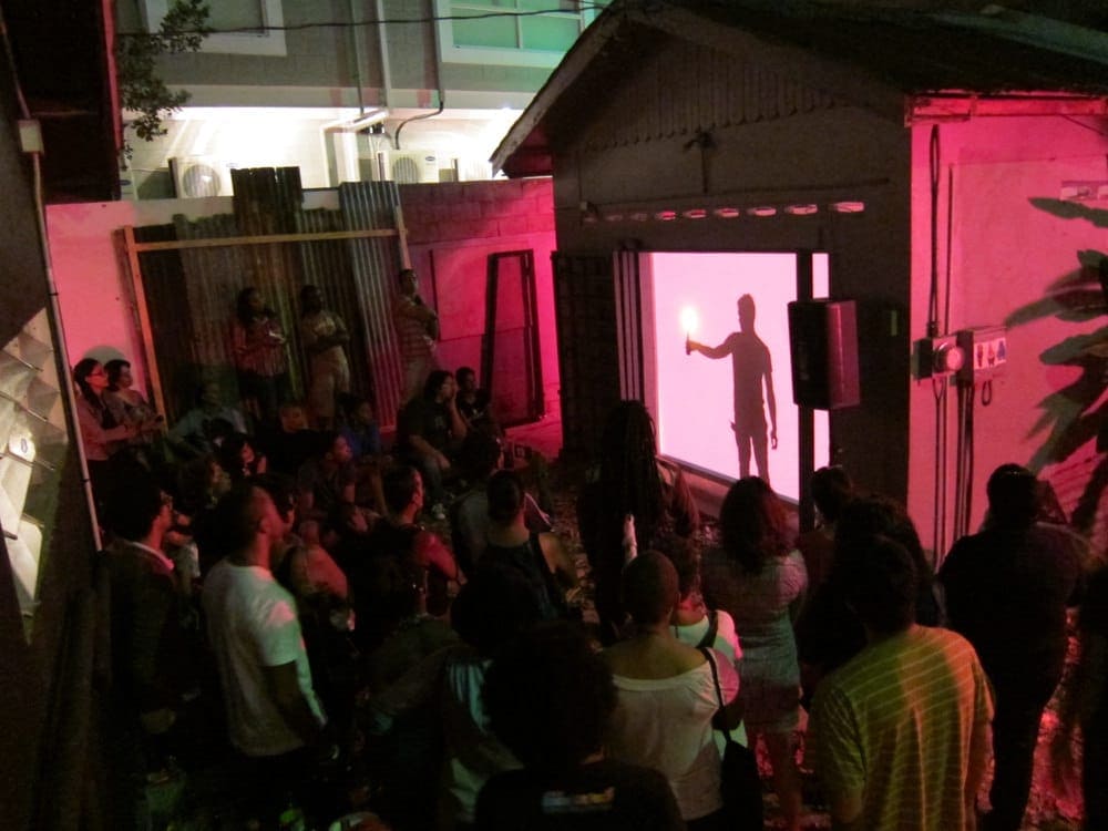 Bagoo performs with a lit flambeau, while the Alice Yard audience looks on.