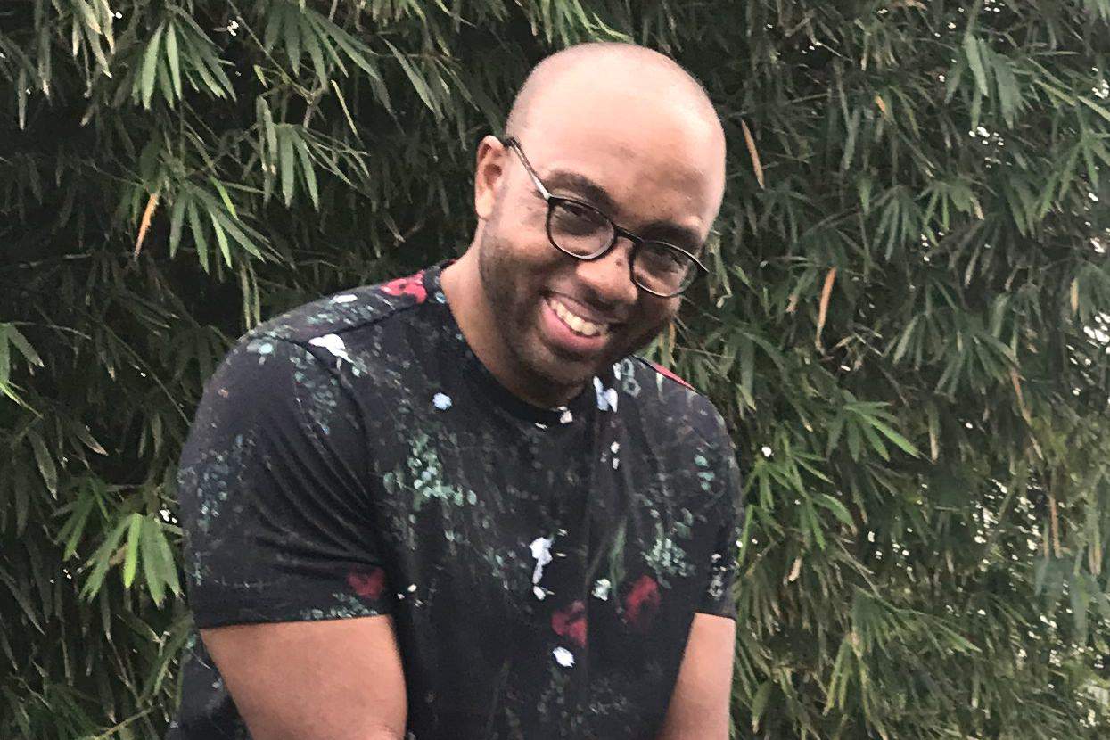 A smiling man with glasses wearing a black floral t-shirt stands in front of bamboo foliage.