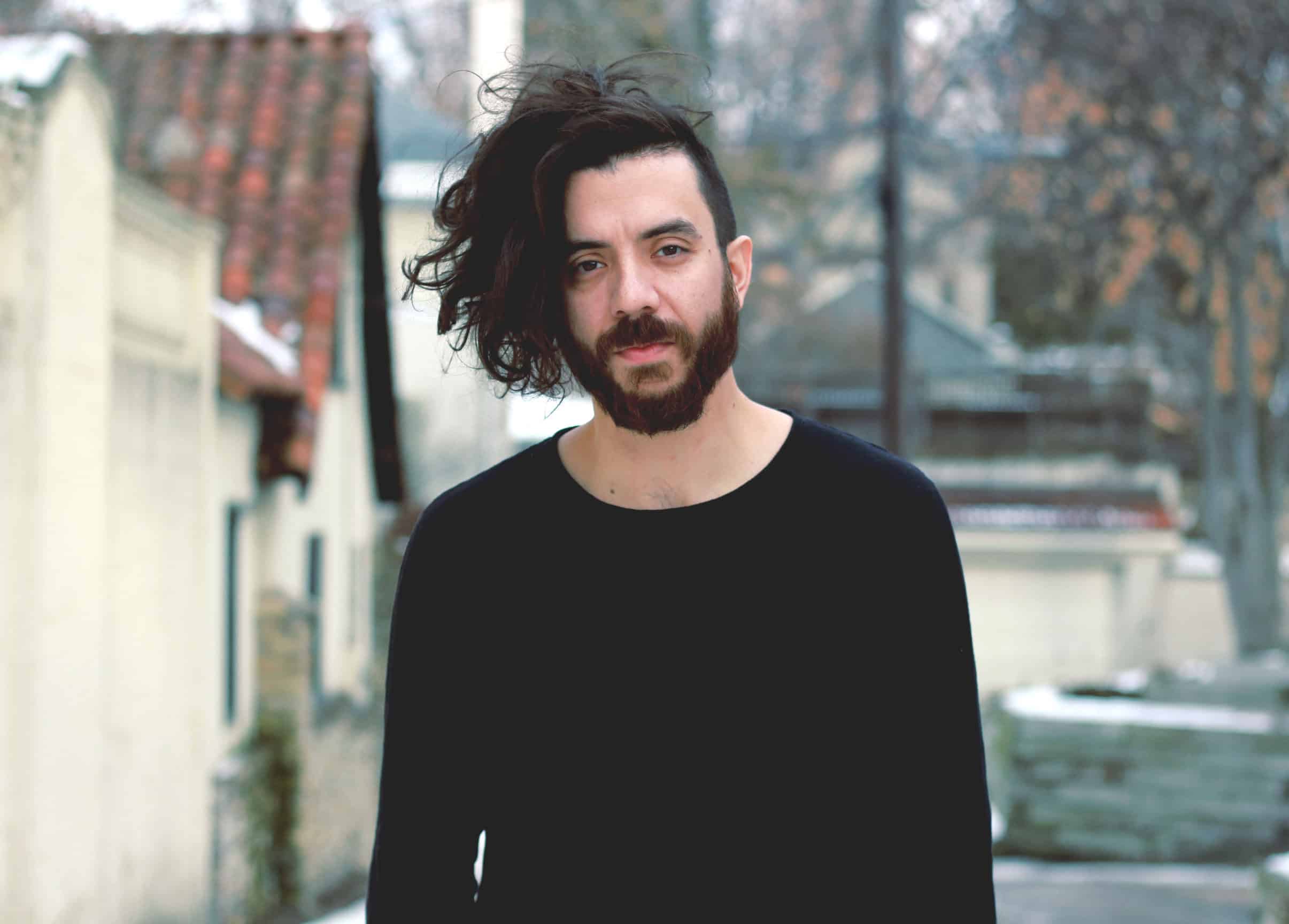 A man with a beard and long, styled hair wearing a black long-sleeved shirt standing outdoors.