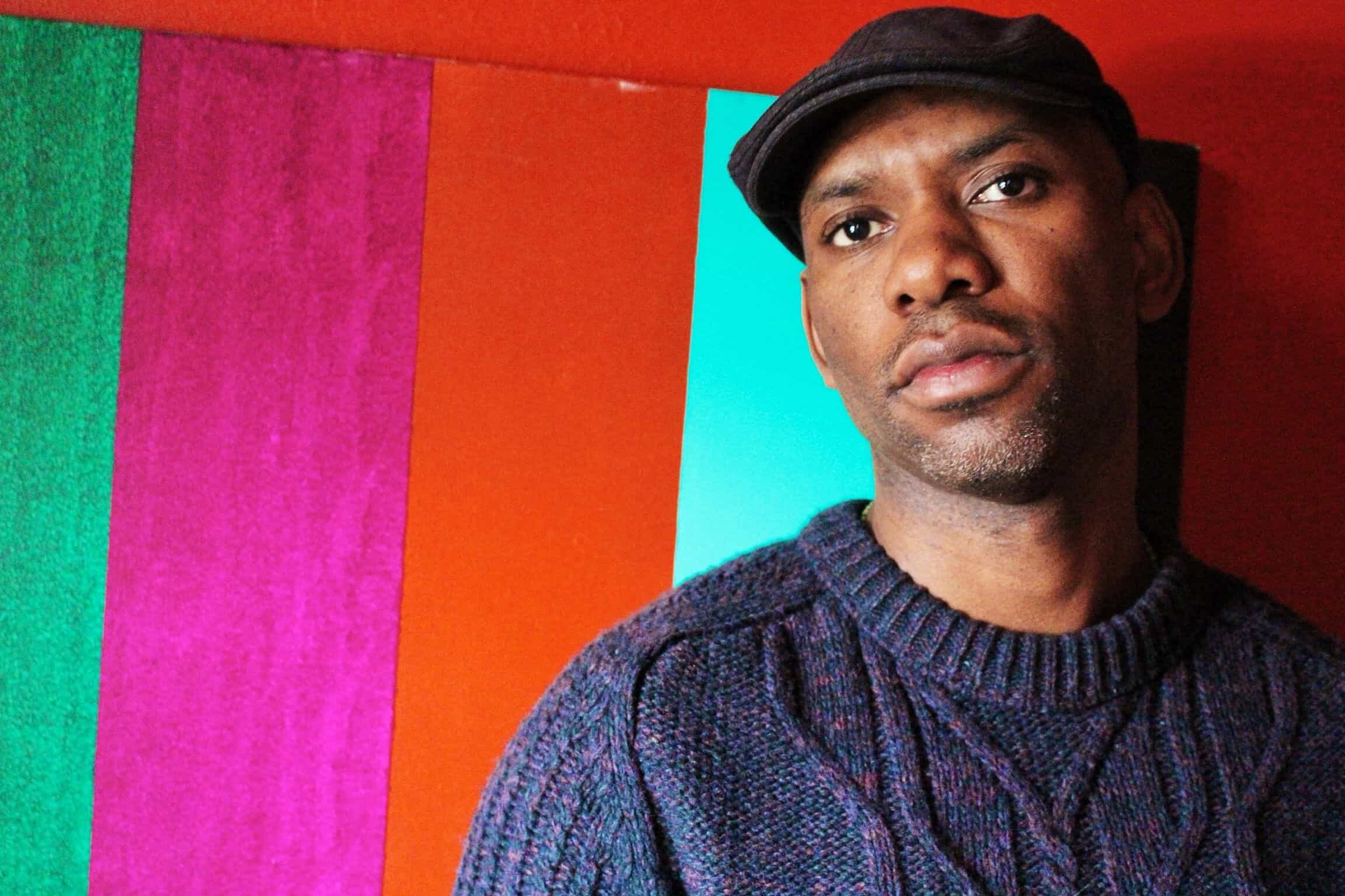Writer and activist Kenyon Farrow wears a flat cap and a dark textured jumper, posing in front of a wall with vertical green, pink, and orange stripes.