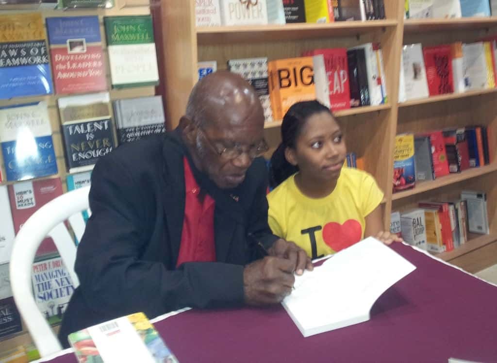 An enthusiastic fan poses alongside Michael Anthony for a photograph, while the author signs one of his books for her.