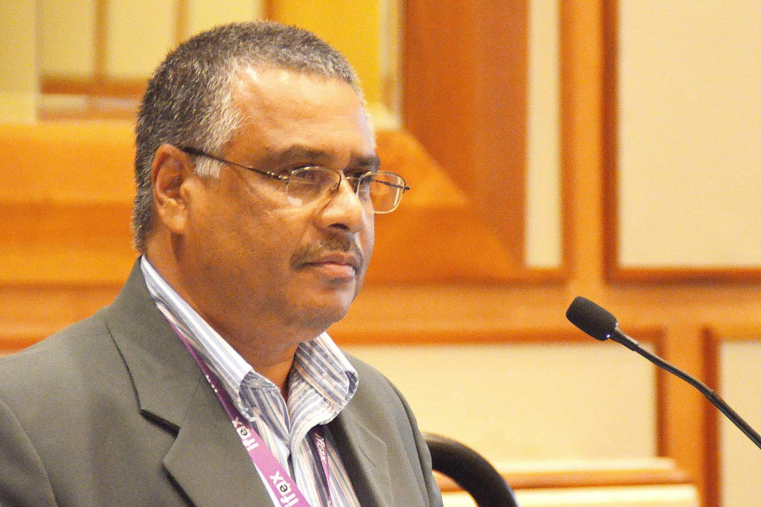 Journalist Wesley Gibbings, wearing glasses and a suit jacket, speaks into a microphone at a conference.