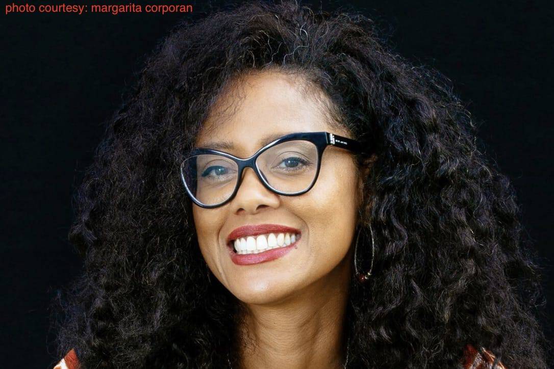 Author and writer Lesley-Ann Brown, a smiling woman with long, curly dark hair and black-framed spectacles.