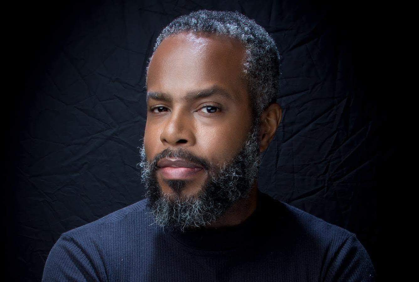 Headshot of Conrad Parris, a middle-aged Black man with greying hair and a beard, wearing a black shirt against a dark background.