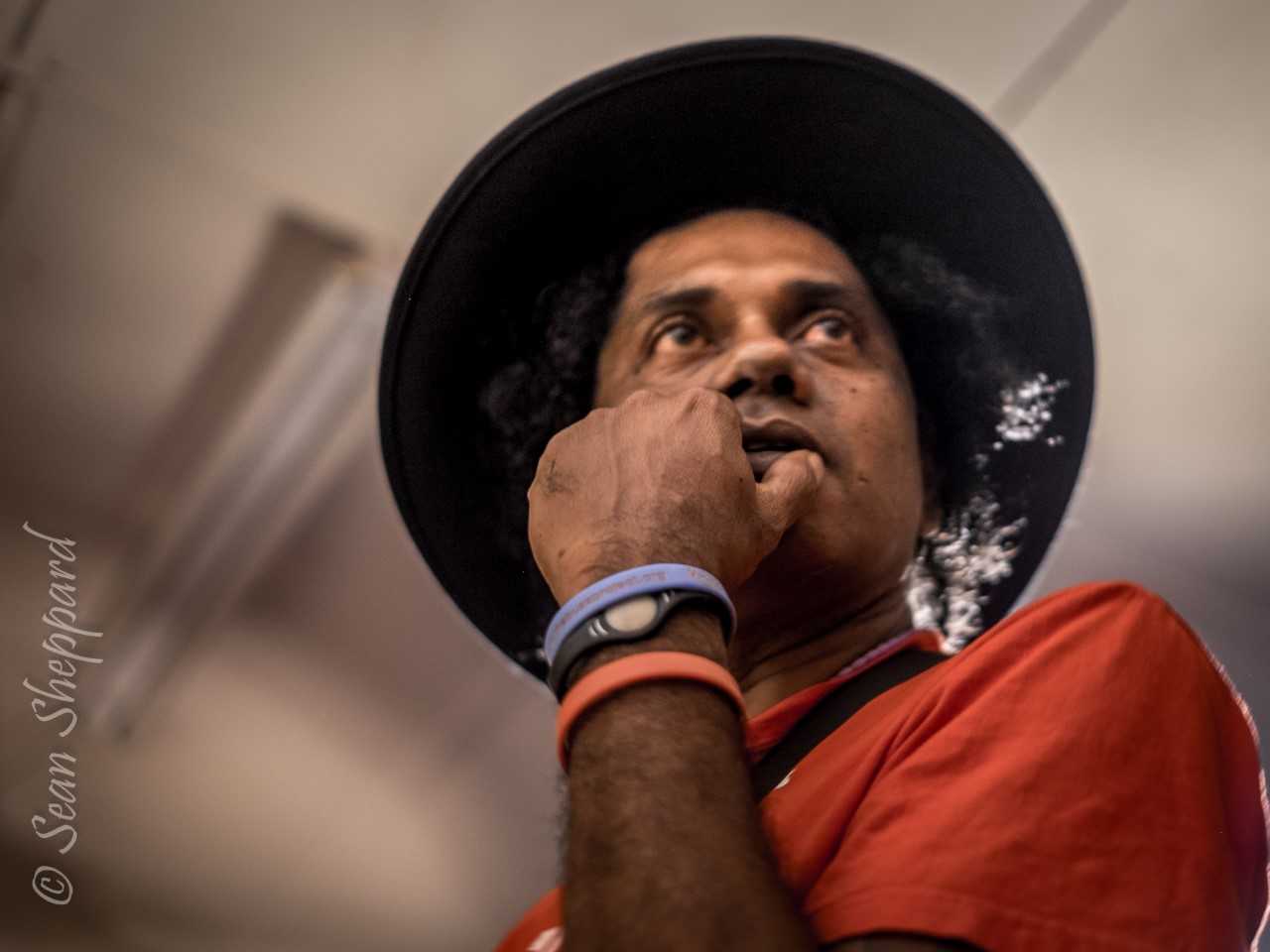 A person wearing a wide-brimmed black hat and an orange shirt looks upwards with a finger near their mouth.