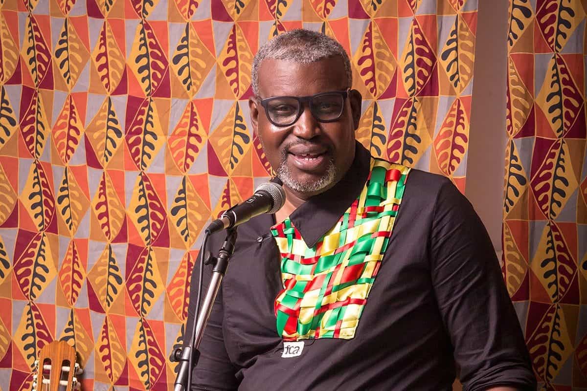 Sterling Henderson, a middle-aged man with grey hair and glasses, speaks into a microphone in front of a patterned fabric backdrop.