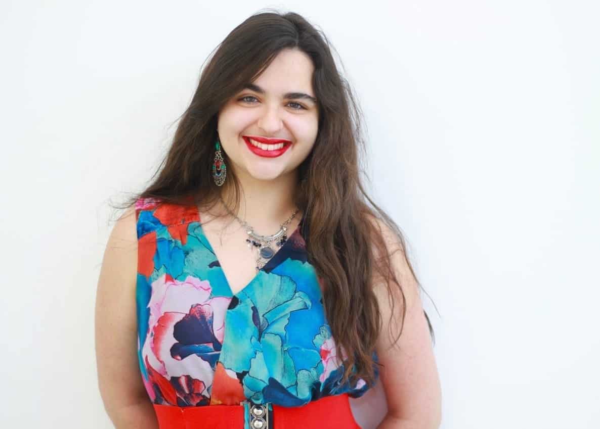 Woman with long brown hair, red lipstick, and colourful floral top stands smiling against a plain white background.