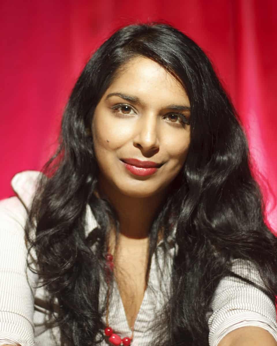 A woman with long dark hair looking directly at the camera against a bright pink background.