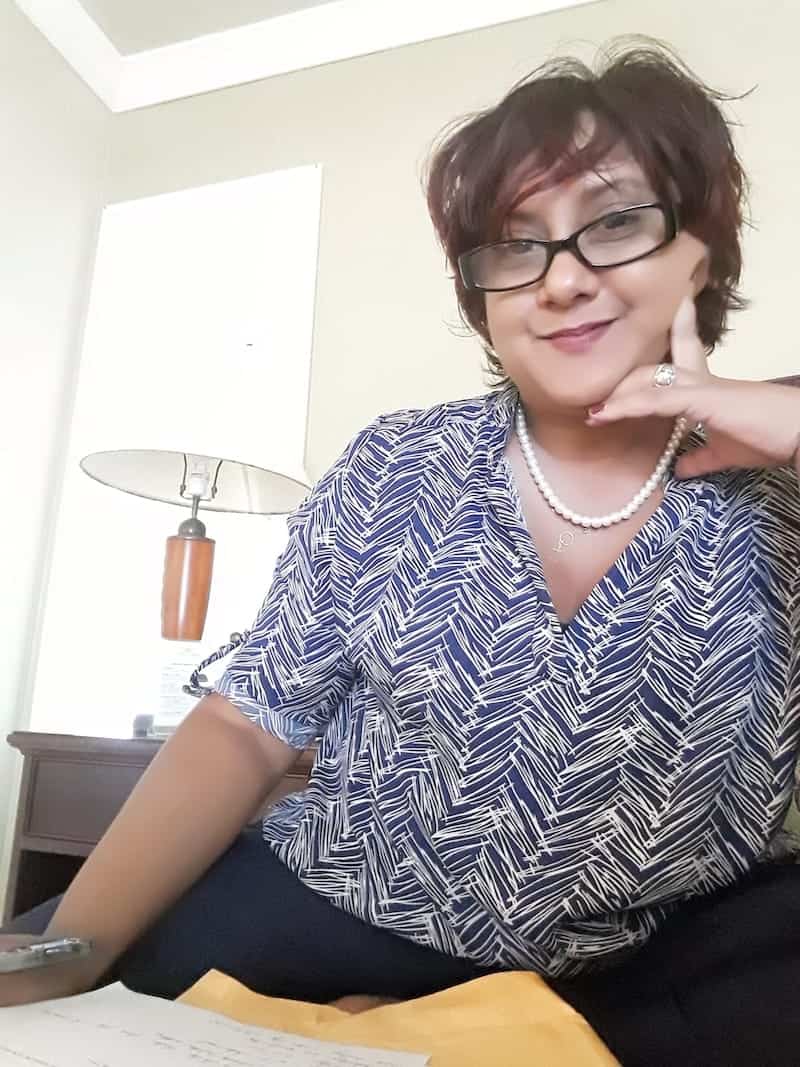 Children’s book author Yolanda T. Marshall, with short hair and glasses, wearing a blue and white patterned shirt and a pearl necklace, rests her chin on her hand at a desk.