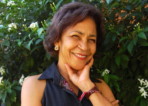A smiling author marketing their book, with shoulder-length dark hair posing in front of a green leafy shrub.