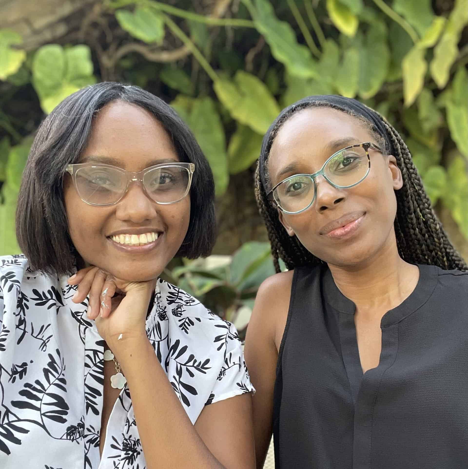 Kernella Berment and another woman wearing glasses pose together in front of green foliage.
