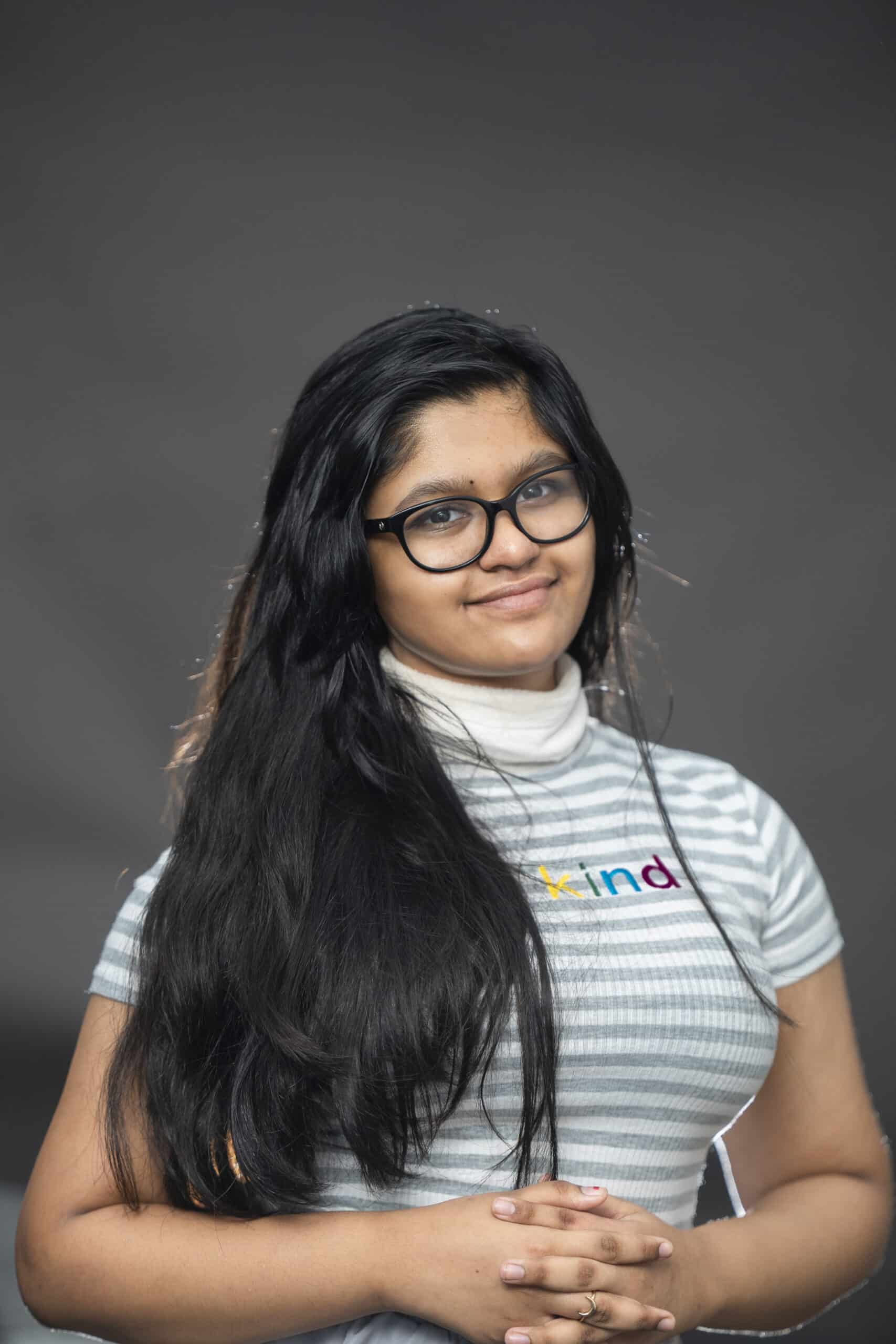 A young woman with long dark hair and glasses wears a grey and white striped shirt with the word “kind” printed on the chest.