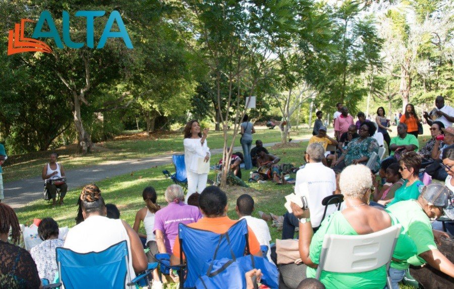 A woman in a white outfit stands and presents online readings to a group of people gathered on chairs and blankets in a park.