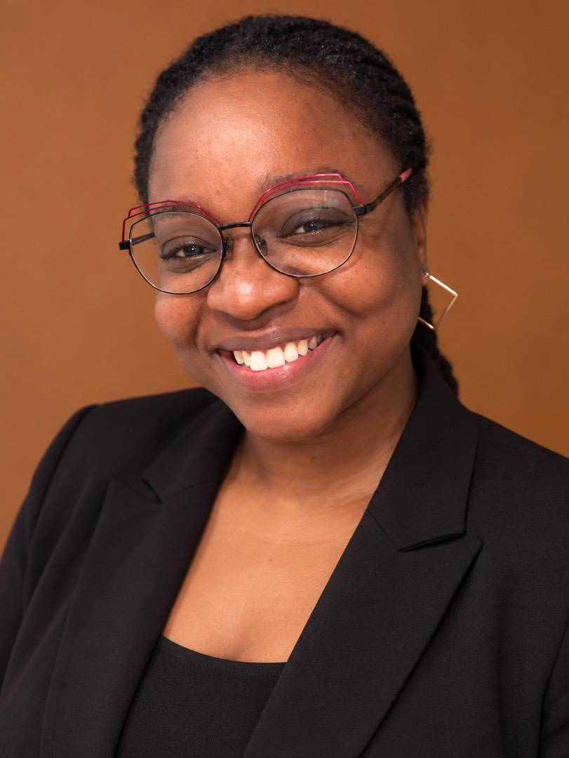 A smiling woman with glasses and a black blazer against a brown background.
