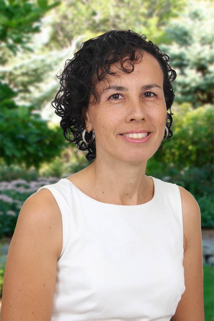 A woman with dark curly hair wearing a white sleeveless top and hoop earrings.