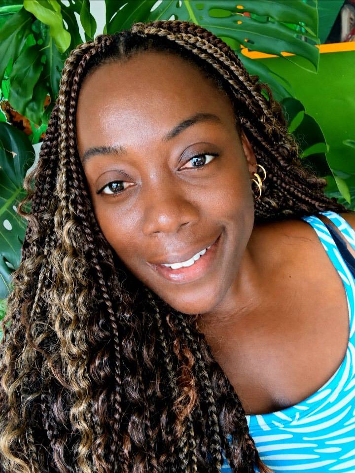 A woman with long, two-toned brown plaited hair smiling in front of green leaves.