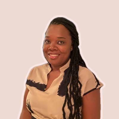 A smiling woman with long locs wearing a tan blouse with a black pattern.