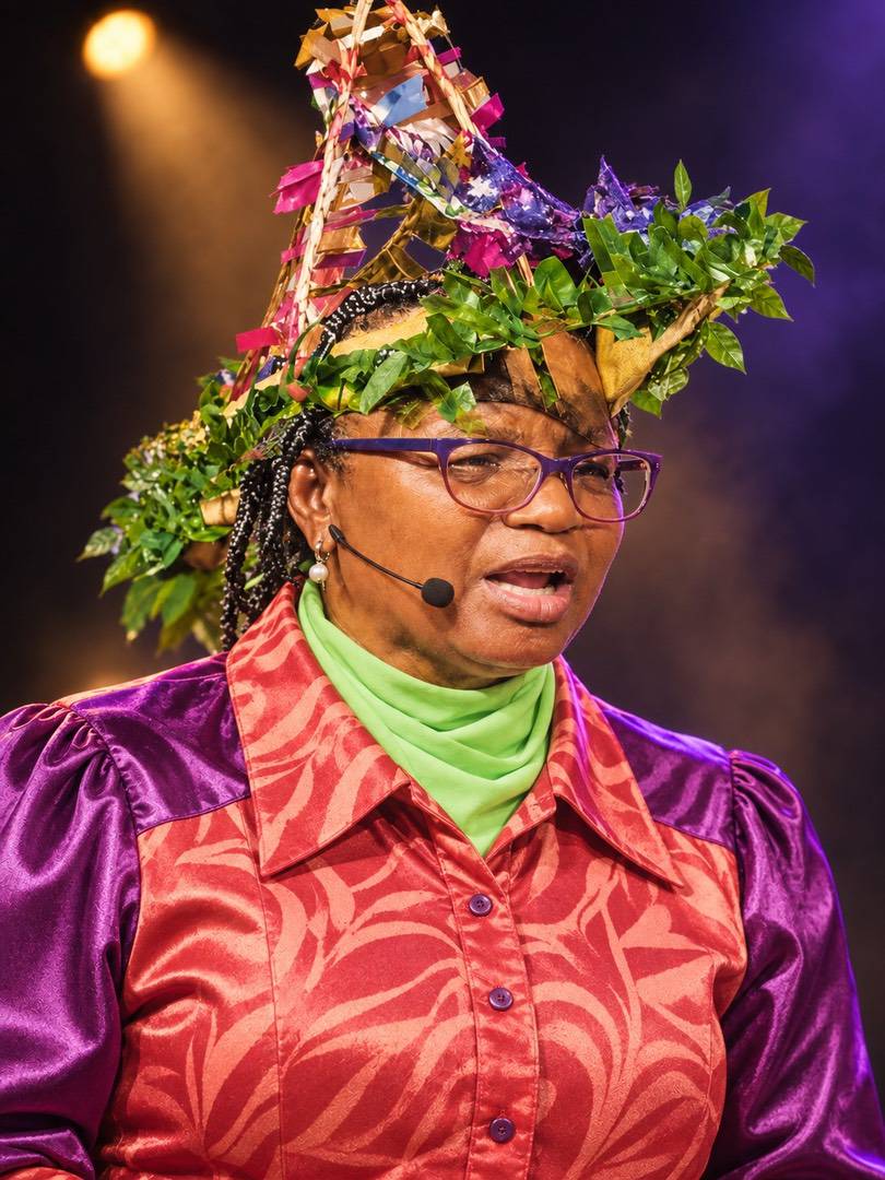 A woman wearing glasses and a headset microphone, dressed in a purple and patterned orange outfit with a decorative, leafy pointed hat.