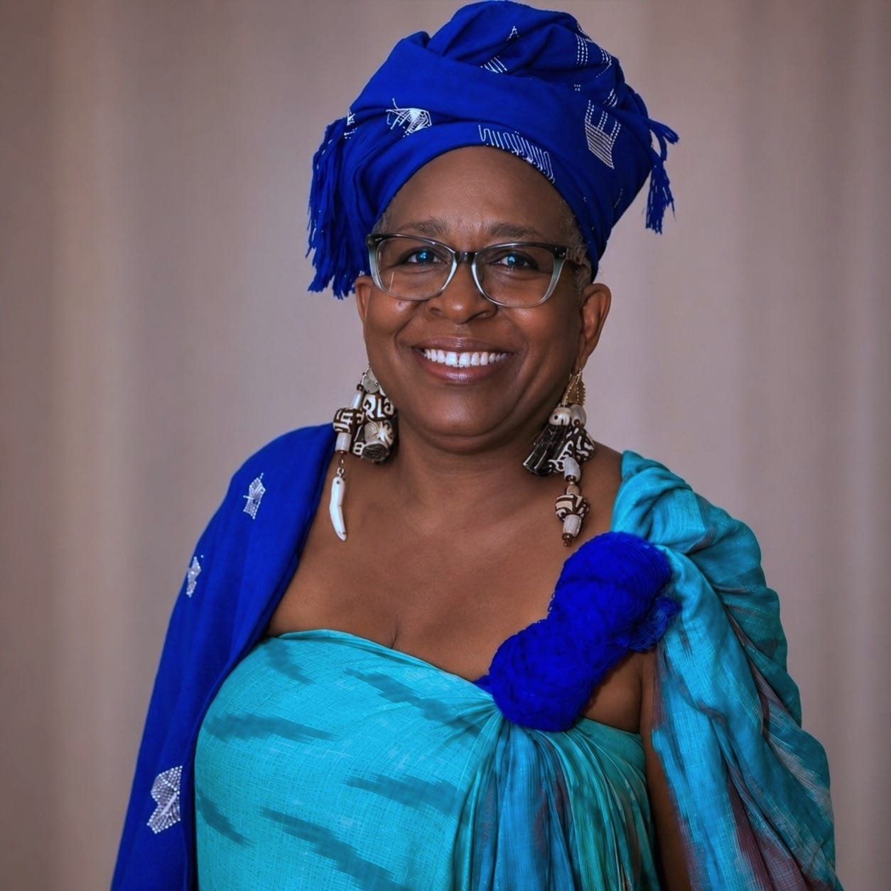 A person smiling, wearing a blue headwrap, a blue dress, and large decorative earrings.
