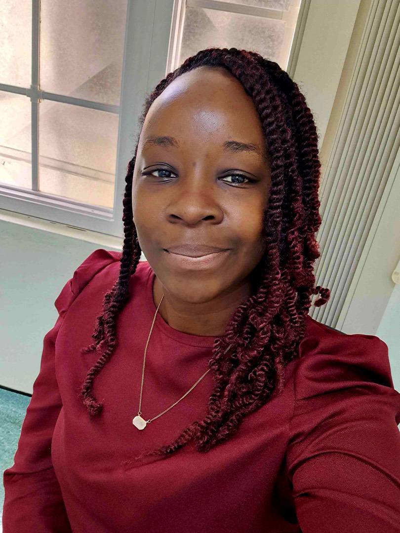 A woman with shoulder-length, dark red plaits wears a maroon long-sleeved top and a gold necklace.