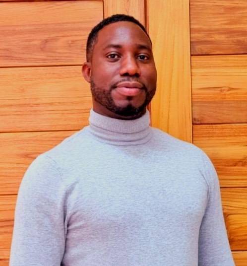 A man with a beard wearing a grey polo-neck jumper standing in front of a wood-panelled wall.