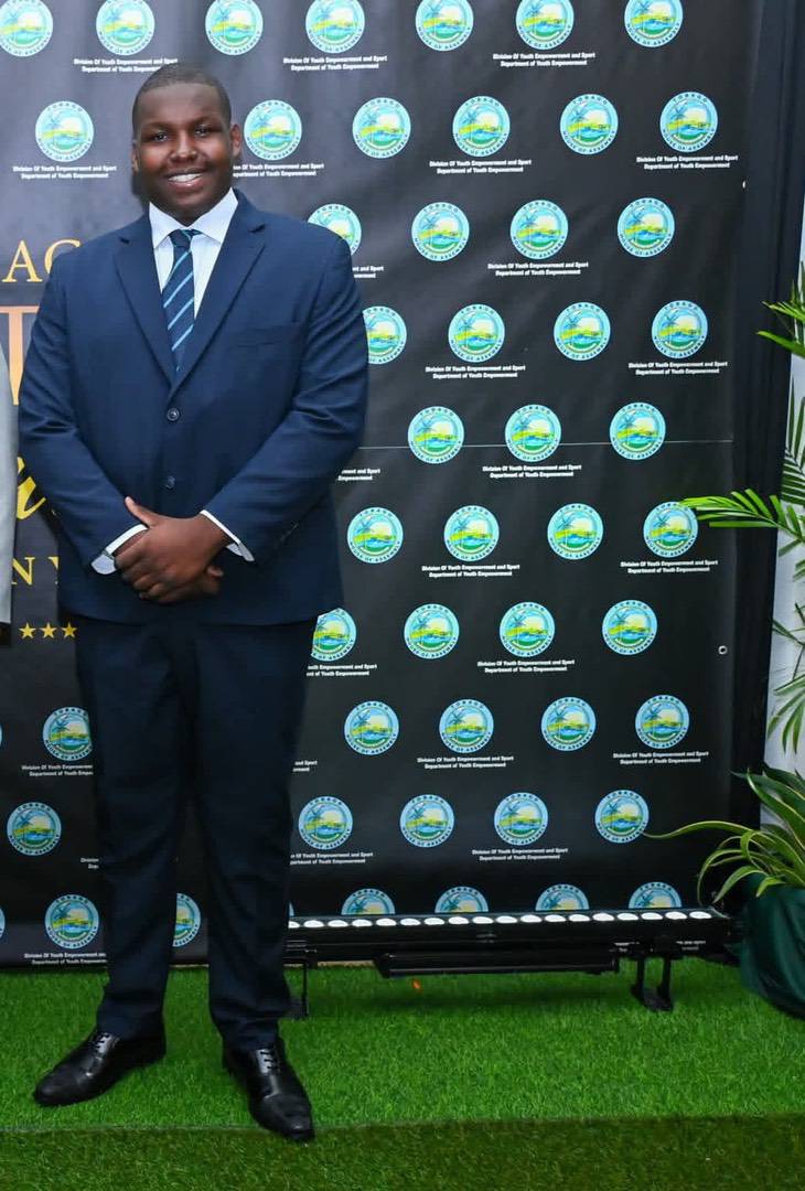 A man in a dark suit and blue tie standing in front of a backdrop featuring a repeating government logo.