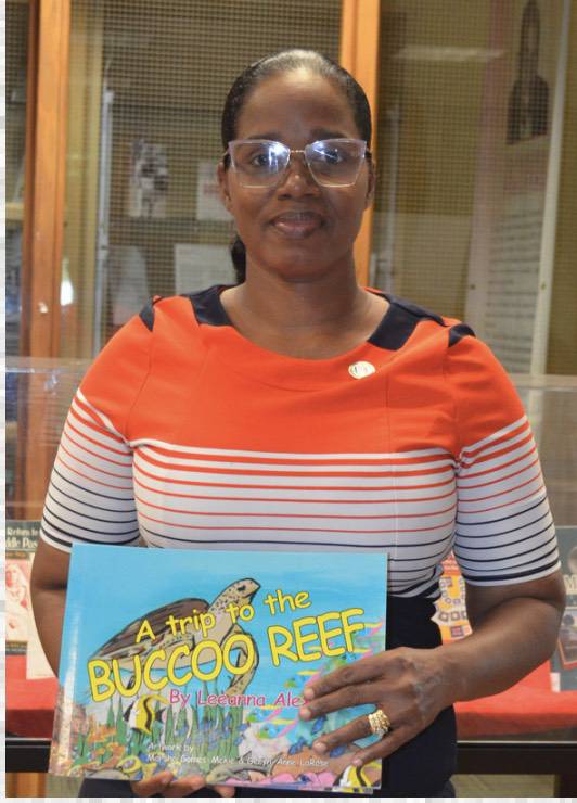 A woman wearing glasses and an orange and white striped shirt holds a children’s book titled “A Trip to the Buccoo Reef” by Leeanna Ale.