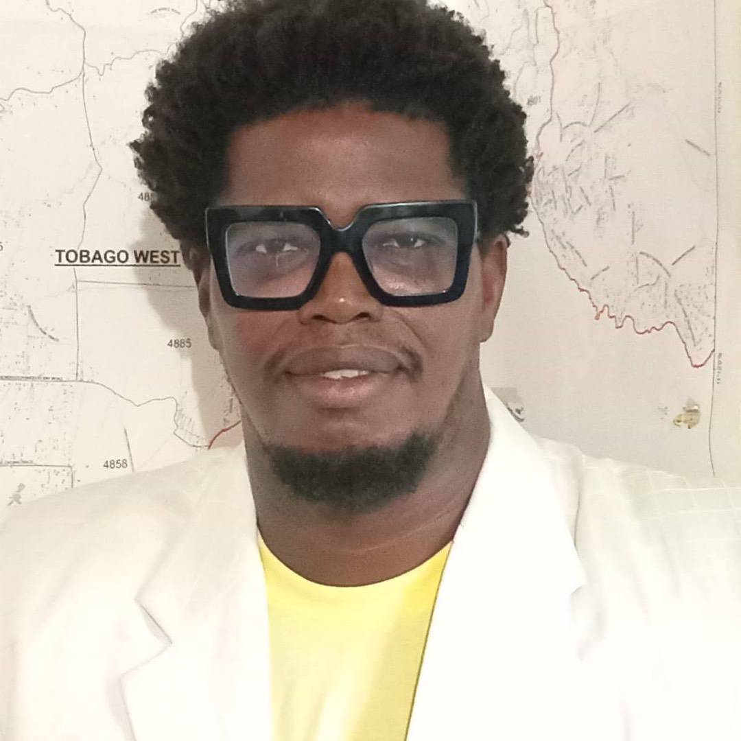 A man with a goatee wearing thick-rimmed glasses, a yellow shirt, and a white blazer, standing in front of a map labelled “Tobago West.”