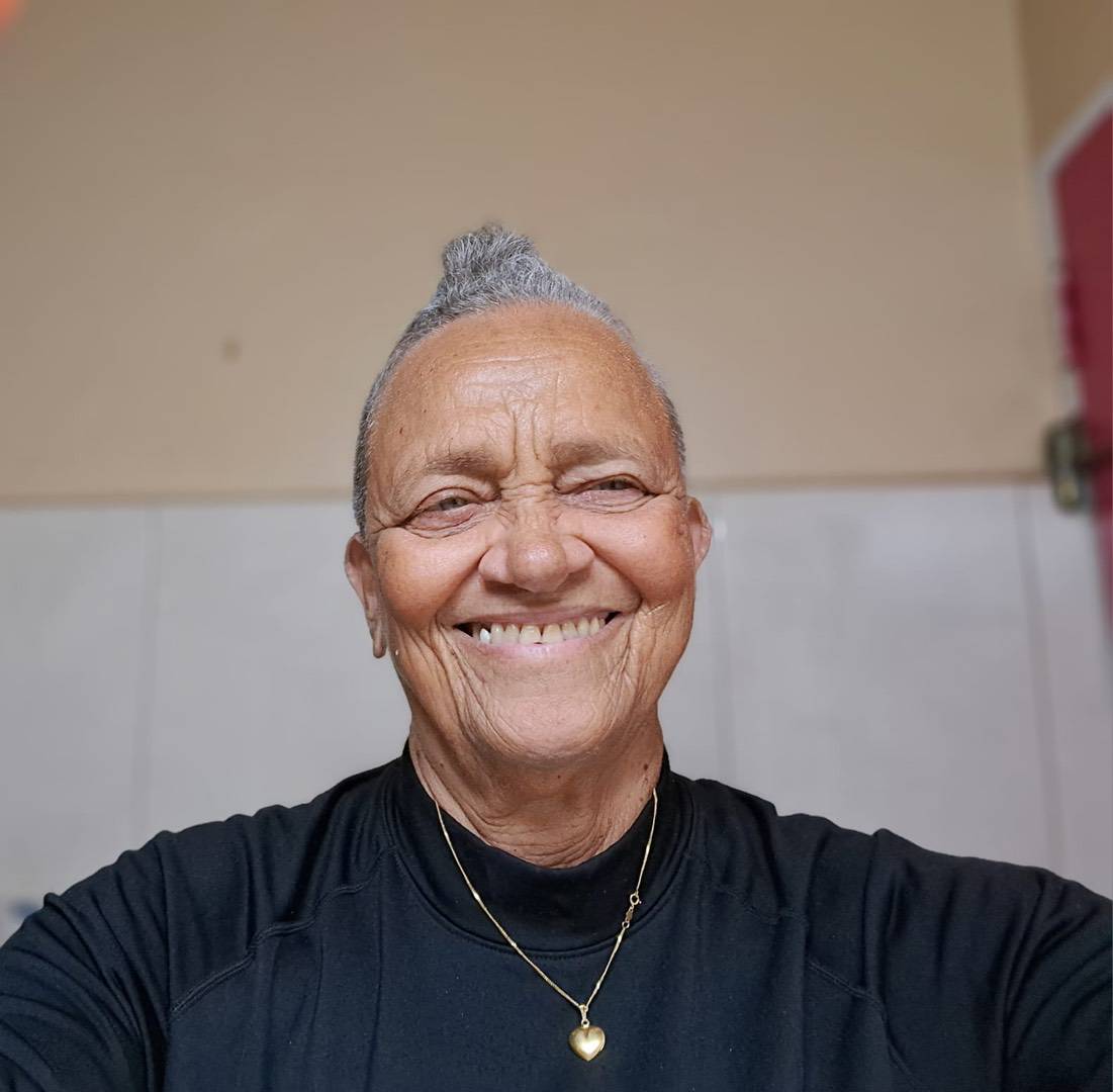 A smiling older woman with grey hair styled in a small topknot, wearing a black shirt and a gold heart pendant necklace.