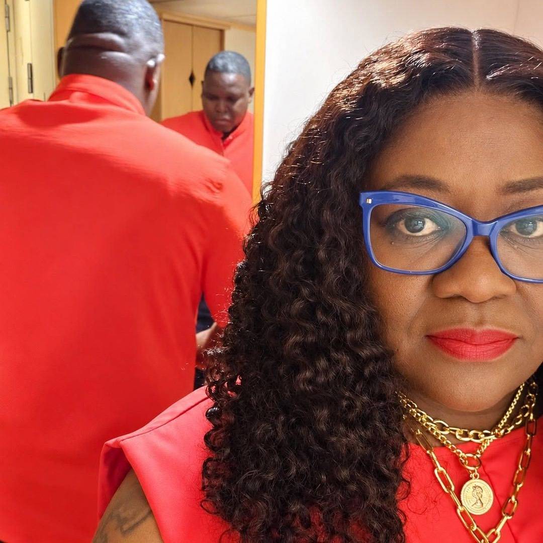 A woman with curly hair, blue cat-eye glasses, and layered gold necklaces, reflected in a mirror behind a man in a red shirt.