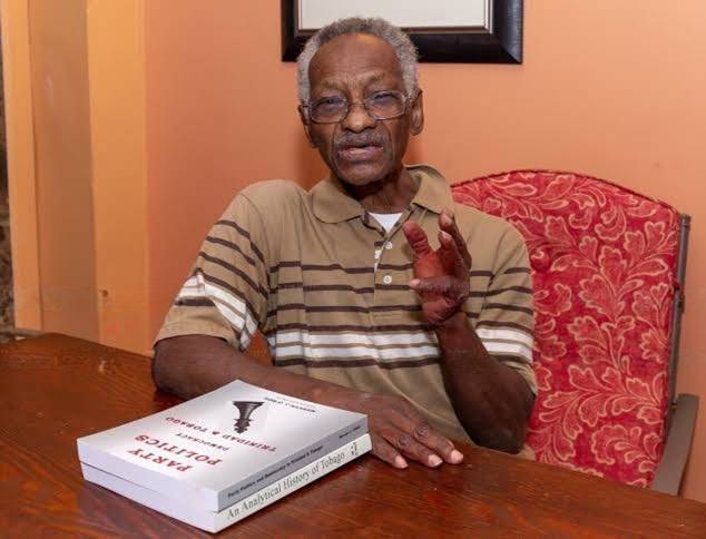 An older man with glasses sits at a table with two books titled “Party Politics in Trinidad and Tobago” and “An Analytical History of Tobago”.