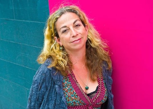 A woman with shoulder-length wavy hair in a patterned top and textured cardigan stands against a wall split between teal and pink in her writing studio.