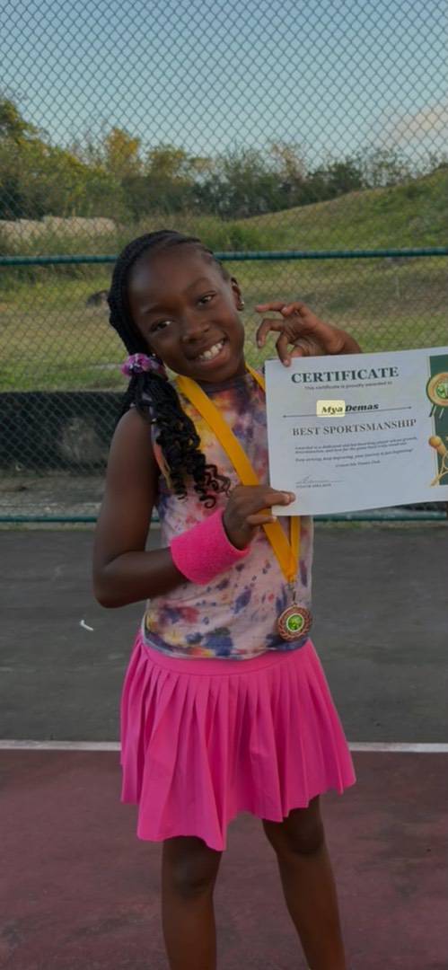 A girl wearing a pink tennis skirt and wristband holds a certificate for “Best Sportsmanship” and wears a medal round her neck.