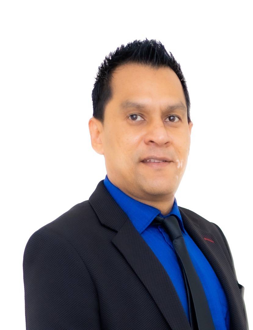 A man wearing a black suit jacket, blue button-up shirt, and dark tie.