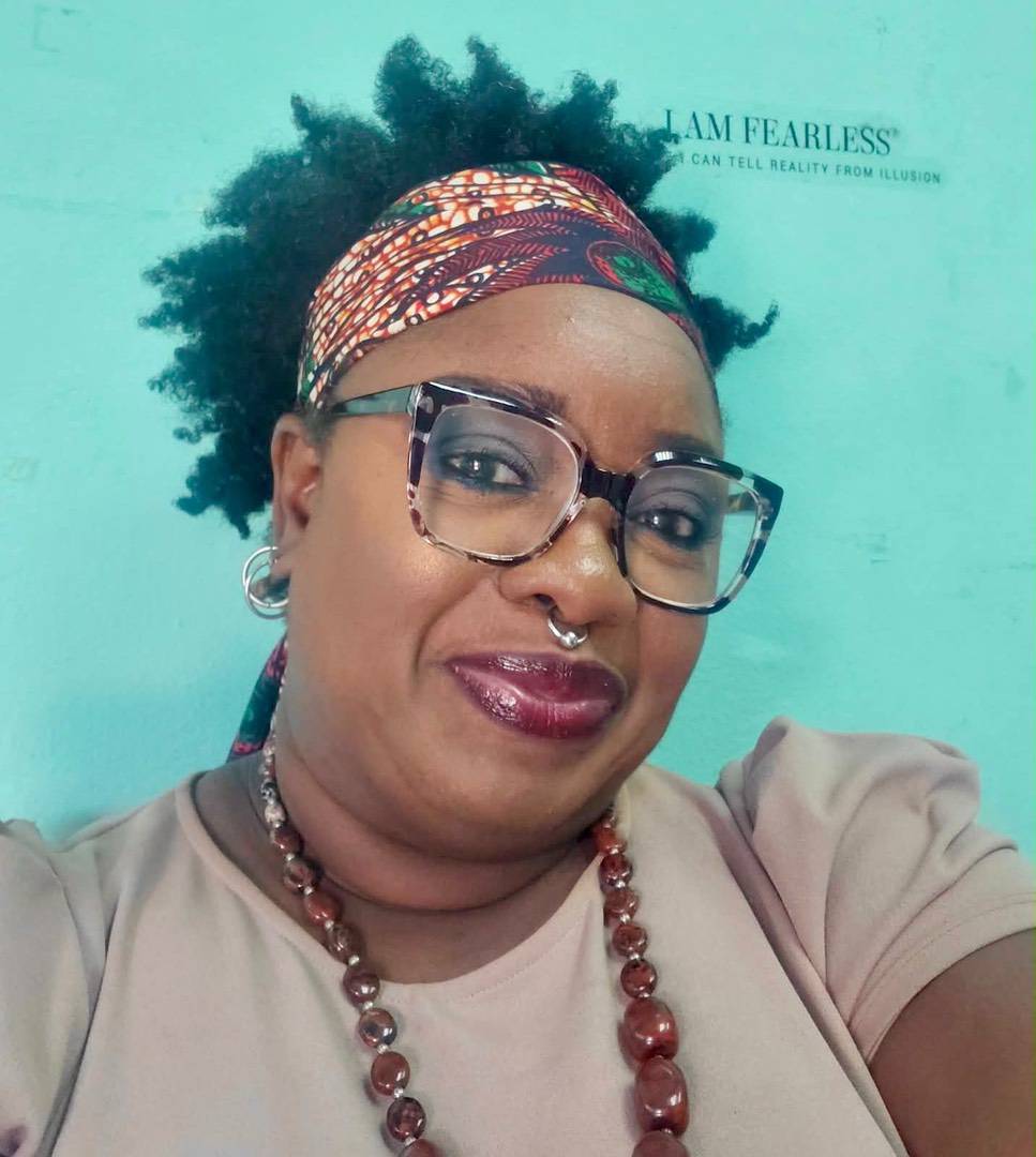 A woman wearing glasses, a patterned headscarf, and a beaded necklace in front of a wall with the text “I AM FEARLESS”.