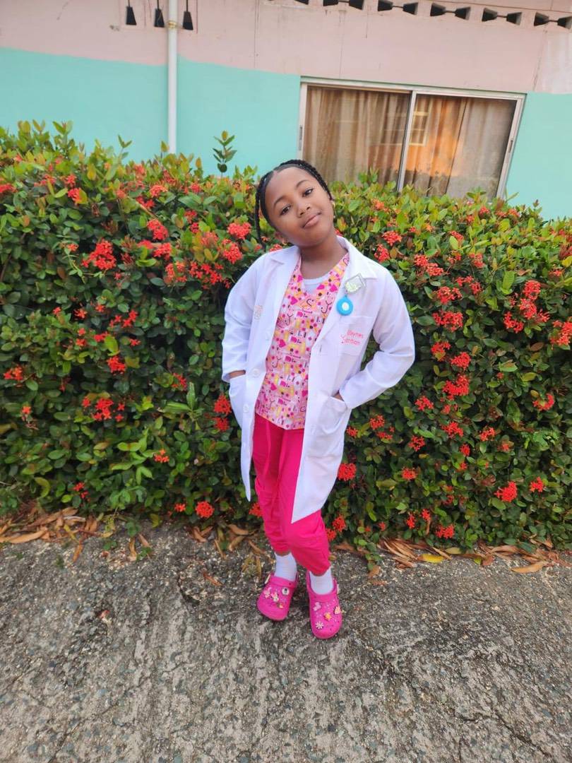 A young girl wearing a white lab coat, patterned scrubs, and pink clogs standing in front of a flowering hedge.