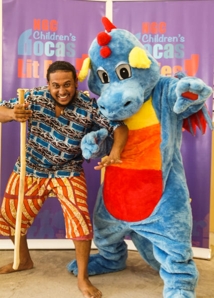 A smiling man holding a wooden staff poses with a blue dragon mascot for a storytelling event in front of an NGC Children’s Bocas Lit Fest backdrop.