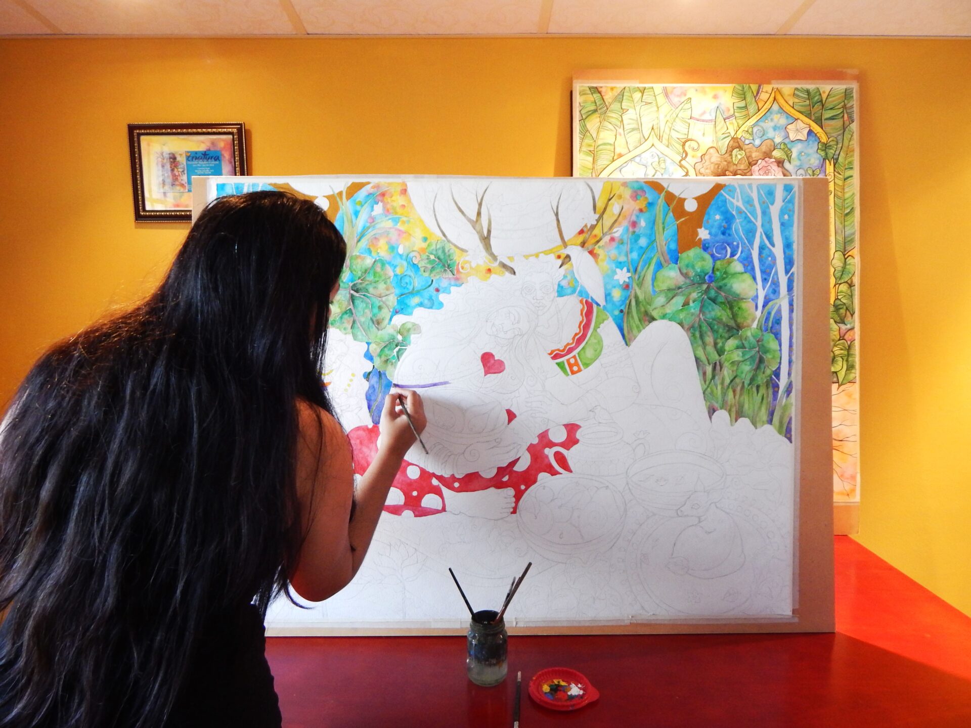 A person with long dark hair painting a large, partially coloured illustration on a canvas during an art class.
